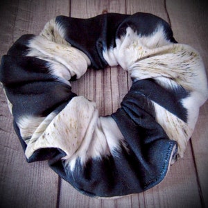 May include: A black and white cow print scrunchie hair tie. The scrunchie is made of a soft, stretchy fabric.
