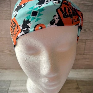 May include: A turquoise headband with a black and white cow print, orange and black geometric shapes, and the text "Mama Tried" repeated on the headband.