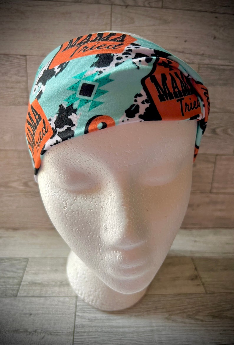 May include: A turquoise headband with a black and white cow print, orange and black geometric shapes, and the text "Mama Tried" repeated on the headband.