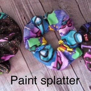 May include: Three colorful hair scrunchies with different patterns. The first scrunchie is black with white speckles. The second scrunchie has a paint splatter design with purple, green, and blue colors. The third scrunchie is black with neon green, pink, and blue designs.