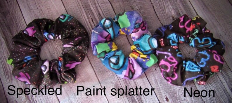 May include: Three colorful hair scrunchies with different patterns. The first scrunchie is black with white speckles. The second scrunchie has a paint splatter design with purple, green, and blue colors. The third scrunchie is black with neon green, pink, and blue designs.
