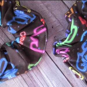 May include: Two black scrunchies with a neon sign print. The scrunchies are made of a soft, stretchy fabric and feature a variety of neon colors, including pink, blue, green, and yellow. The scrunchies are perfect for adding a pop of color to any outfit.
