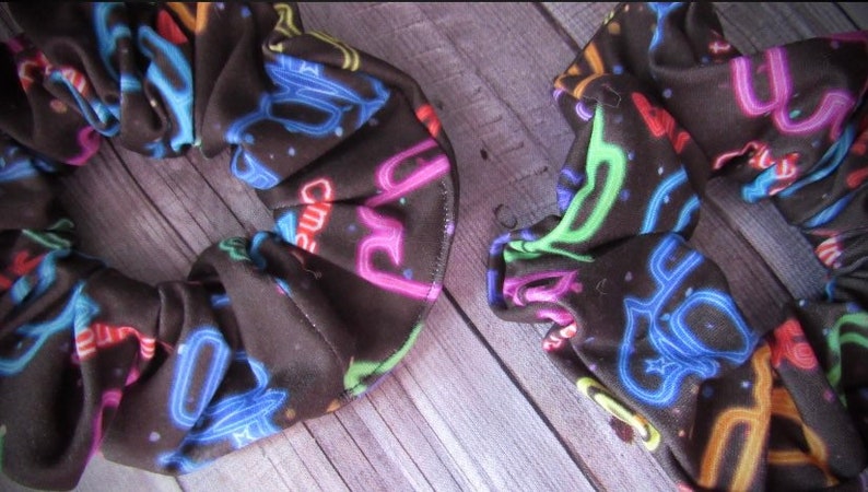 May include: Two black scrunchies with a neon sign print. The scrunchies are made of a soft, stretchy fabric and feature a variety of neon colors, including pink, blue, green, and yellow. The scrunchies are perfect for adding a pop of color to any outfit.