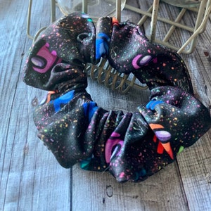 May include: Black scrunchie with a colorful print of the characters from the video game Among Us. The scrunchie is made of a soft, stretchy fabric.