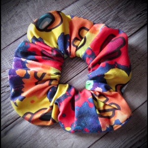May include: A colorful scrunchie with a tie-dye pattern featuring yellow, red, blue, and black. The scrunchie has a repeating pattern of cartoon characters.