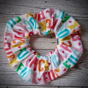 May include: A white fabric hair scrunchie with a colorful alphabet print and small pink hearts.