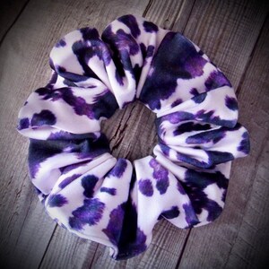 May include: A scrunchie with a white and purple spotted pattern. The fabric is gathered and bunched, creating a voluminous, circular shape. The scrunchie is designed to hold hair in a ponytail or bun.