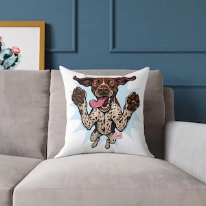 May include: Square white cushion with a cartoon dog illustration. The dog has brown and white spots, a pink tongue, and raised paws, set against a light blue burst. The cushion is on a grey sofa.
