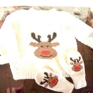 Puede incluir: Un conjunto de suéter y mitones de punto blanco. El suéter y los mitones presentan un diseño de reno con cuernos marrones, una nariz roja y una cara marrón. El conjunto es probablemente para un bebé o un niño pequeño.