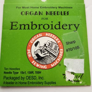 May include: Green packet of ORGAN NEEDLES for embroidery. The packet features the text "ORGAN NEEDLES FOR Embroidery" and a circular logo with a woman using a sewing machine. The packet also includes the text "Sharp #70/10S".