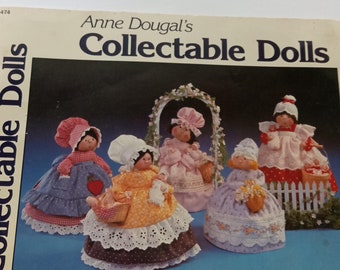 May include: A book titled "Anne Dougals's Collectible Dolls" with a cover image of five handmade dolls dressed in colorful clothing. The dolls are arranged in front of a white picket fence and a floral archway.