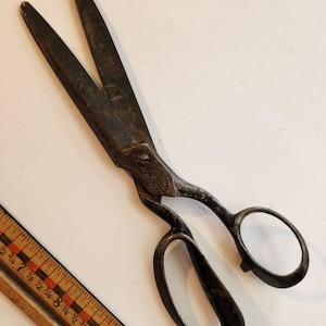 以下が含まれることがあります： 暗い、古びた仕上げの、ヴィンテージの大きな金属製のハサミ。ハサミには大きな指穴と長い刃が付いています。黒と赤の目盛りの木製定規も画像にあります。