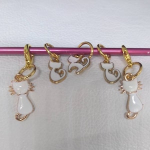 May include: Four gold-coloured cat-shaped stitch markers with white enamel. The cats are in different poses, including sitting, standing, and curled up.