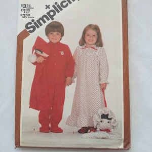 May include: Simplicity sewing pattern number 5779 for a toddler's sleepwear set. The pattern includes a red sleepsuit with a white trim and a white nightgown with a red trim. The pattern is for a toddler girl and boy.