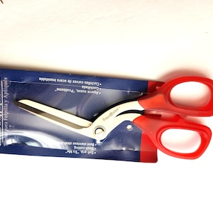 May include: Fons & Porter chenille and appliqué scissors with red handles, presented on a blue and red package. The scissors feature a curved blade, ideal for felting and appliqué. The packaging includes text in English and Spanish.