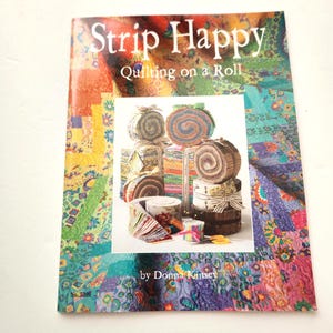 May include: A book titled "Strip Happy Quilting on a Roll" by Donna Kinsey. The cover features a colorful patchwork design and an image of rolled fabric strips. The book is surrounded by a white background.
