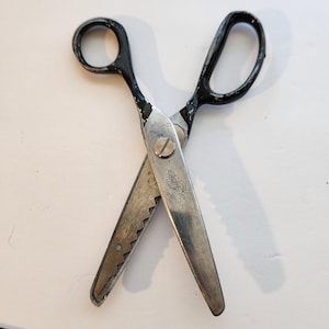 May include: Vintage metal scissors with black handles. The blades are silver with a serrated edge on one side. The scissors are open, displaying the blades and the screw that holds them together. The handles show signs of wear.