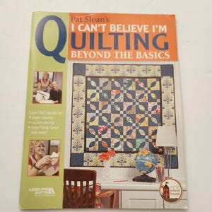 Op de afbeelding: Een instructieboek met zachte kaft getiteld Pat Sloan's I CAN'T BELIEVE I'M QUILTING BEYOND THE BASICS. De kaft toont een grote blauwe letter Q, een kleurrijke quilt met patronen en kleine foto's van een persoon die naait en met stof werkt.