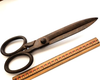 May include: Large, antique metal scissors with dark brown handles and blades. A wooden ruler with red and black markings is next to the scissors. The scissors are approximately 12 inches long.