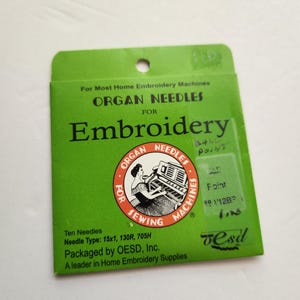 Peut inclure: Un emballage rectangulaire vert contenant dix aiguilles à broder Organ pour machines domestiques. L'emballage présente un logo circulaire avec une illustration de machine à coudre, le texte For Most Home Embroidery Machines et le type 15x1, 130R, 705H.