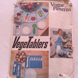 Op de afbeelding: Een vintage Vogue Patterns naaipatroon voor een set "Vegetablers" placemats en servetten. Het patroon heeft een blauw-wit geruit tafelkleed met placemats en servetten in de vorm van groenten. Het patroonnummer is 692.