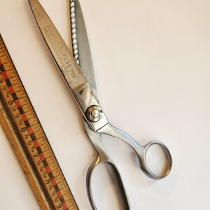 May include: Vintage metal sewing shears with the text "Rivington Shears" and a wooden ruler with red and black markings. The shears have a silver finish and are open, displaying the blades.