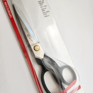 Könnte beinhalten: Superfection Classic Schere in einer transparenten Kunststoffverpackung. Die Schere hat schwarze Griffe und silberne Klingen. Ein rotes Band ist um die Griffe gewickelt. Der Text "Superfection Classic" ist rot.
