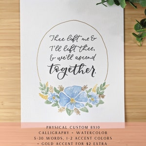 May include: A white rectangular print featuring black calligraphy text that reads, Thee lift me & I'll lift thee, & we'll ascend together. The text is enclosed in a thin gold oval frame with a watercolor floral arrangement of blue flowers and green leaves at the base.