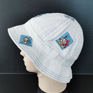 May include: White bucket hat with blue stitching and cartoon character patches. The hat has a brim and is made of a soft, textured fabric. Patches feature Donald Duck and Goofy.