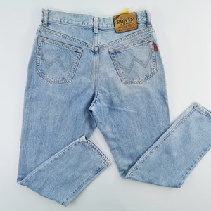 May include: Light blue denim jeans with a classic five-pocket design. The jeans feature a straight leg cut and a faded wash. The back pockets have a distinctive stitched design. The Edwin brand label is visible on the back waistband.