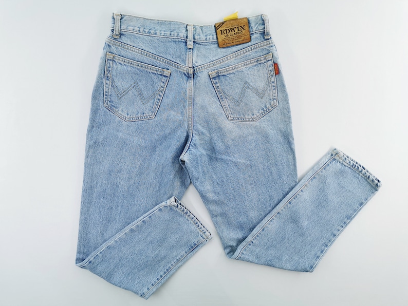 May include: Light blue denim jeans with a classic five-pocket design. The jeans feature a straight leg cut and a faded wash. The back pockets have a distinctive stitched design. The Edwin brand label is visible on the back waistband.