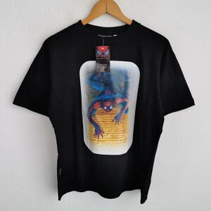 Pode incluir: Camiseta preta com um gr&aacute;fico do Homem-Aranha em uma moldura retangular. A imagem mostra o super-her&oacute;i em uma pose din&acirc;mica contra uma parede de tijolos. A camiseta tem uma etiqueta presa &agrave; gola.