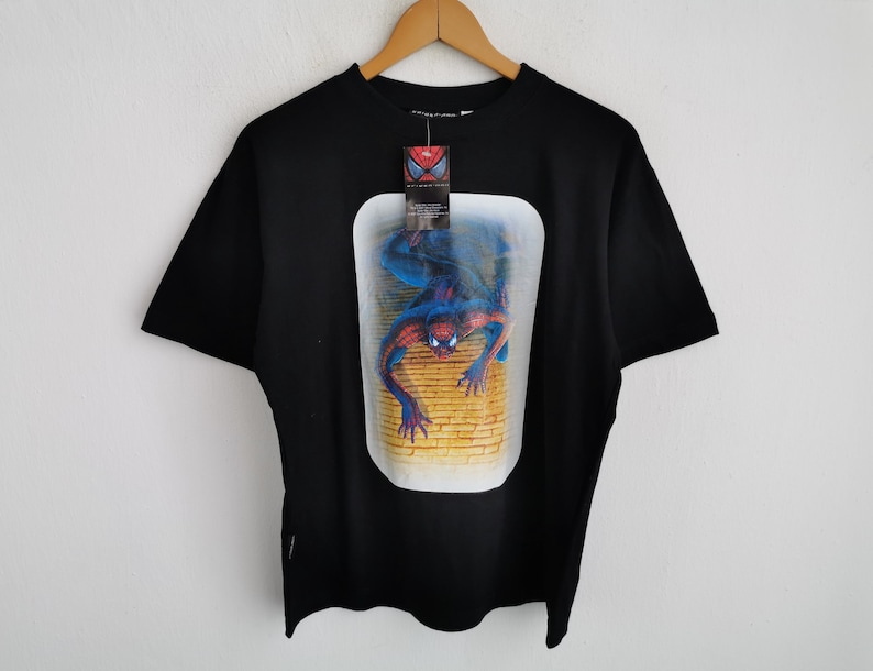 Pode incluir: Camiseta preta com um gr&aacute;fico do Homem-Aranha em uma moldura retangular. A imagem mostra o super-her&oacute;i em uma pose din&acirc;mica contra uma parede de tijolos. A camiseta tem uma etiqueta presa &agrave; gola.