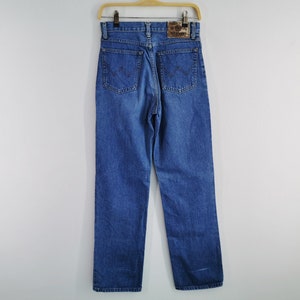 May include: Blue denim jeans with a straight leg design, hanging on a wooden hanger. The jeans feature classic five-pocket styling, a zip fly, and a leather patch on the back waistband. The Wrangler logo is visible on the patch.