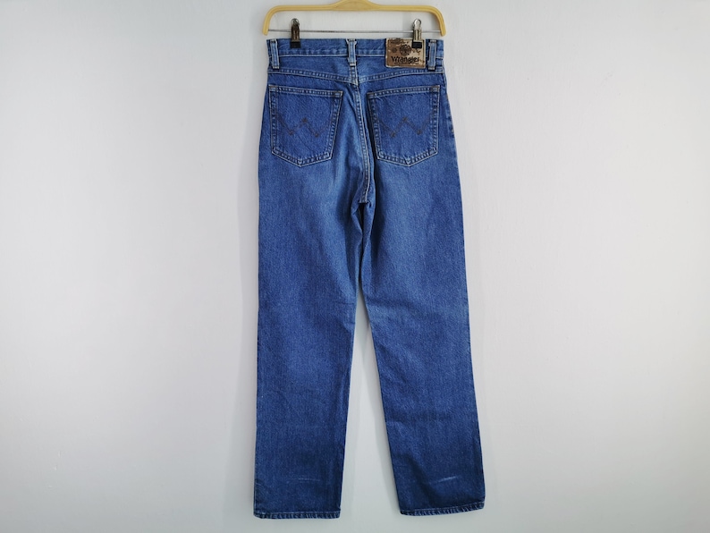 May include: Blue denim jeans with a straight leg design, hanging on a wooden hanger. The jeans feature classic five-pocket styling, a zip fly, and a leather patch on the back waistband. The Wrangler logo is visible on the patch.