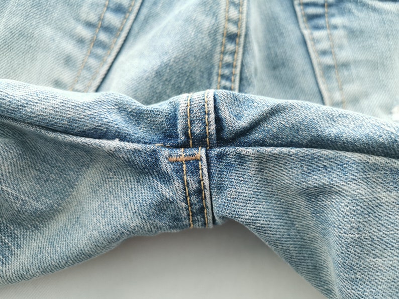 May include: Close-up of light blue denim jeans, showcasing the detailed stitching and fabric texture. The image highlights the seams and the worn appearance of the denim, emphasizing the garment's construction and style.