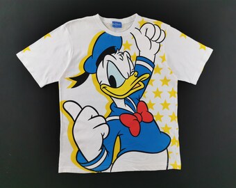 Puede incluir: Una camiseta blanca de manga corta con un gran gráfico de Donald Duck con un sombrero de marinero azul y una pajarita roja. El personaje gesticula con ambas manos, rodeado por un patrón de estrellas amarillas en el lado derecho.