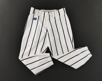 Puede incluir: Pantalones de béisbol blancos con rayas verticales azul marino, cierre de botón, trabillas para cinturón y dobladillo elástico en los tobillos. Un pequeño parche rectangular con una letra M azul claro sobre fondo negro se encuentra en la cadera derecha.