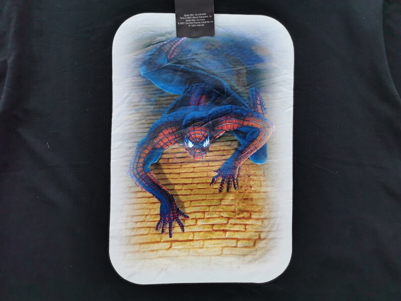 Pode incluir: Um gr&aacute;fico retangular do Homem-Aranha sobre um fundo preto. A imagem apresenta o Homem-Aranha numa pose din&acirc;mica contra uma parede de tijolos. O super-her&oacute;i &eacute; retratado no seu cl&aacute;ssico fato vermelho e azul.