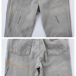 May include: Light gray denim jeans with a classic five-pocket design. The jeans show signs of wear and tear, with visible staining and a few small tears. The back pockets feature a stitched design, and a brown leather patch is visible.