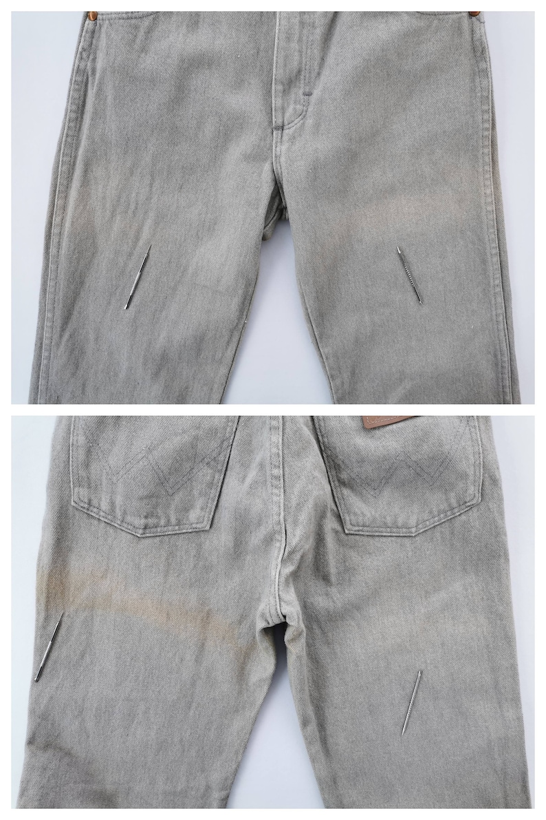 May include: Light gray denim jeans with a classic five-pocket design. The jeans show signs of wear and tear, with visible staining and a few small tears. The back pockets feature a stitched design, and a brown leather patch is visible.