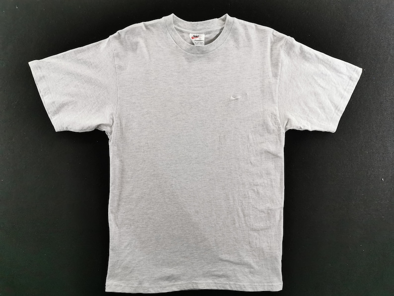 May include: A light gray t-shirt with a crew neck and short sleeves. The Nike logo is embroidered in white on the upper left chest. The shirt is made of a soft, likely cotton, material.