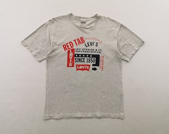 Puede incluir: Una camiseta de manga corta en color gris jaspeado claro con un estampado gráfico que incluye el texto RED TAB LEVI'S, LEVI STRAUSS & CO. SAN FRANCISCO CAL., SINCE 1850 y WE NEVER GOES OUT OF STYLE, junto a un logo rojo de Levi's y cinco estrellas.