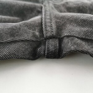 May include: Close-up of the waistband and fly of a pair of dark gray denim jeans. The fabric has a textured appearance with visible diagonal twill lines. The stitching is visible, and the jeans appear to be new.