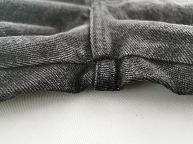 May include: Close-up of the waistband and fly of a pair of dark gray denim jeans. The fabric has a textured appearance with visible diagonal twill lines. The stitching is visible, and the jeans appear to be new.