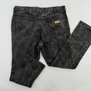 May include: A pair of black denim jeans with a faded, textured appearance. The jeans feature a classic five-pocket design, belt loops, and a Wrangler brand patch on the back pocket. The jeans are laid flat on a white surface.