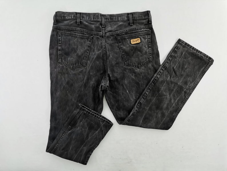 May include: A pair of black denim jeans with a faded, textured appearance. The jeans feature a classic five-pocket design, belt loops, and a Wrangler brand patch on the back pocket. The jeans are laid flat on a white surface.