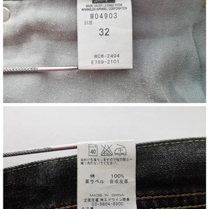 May include: Close-up of the inside of a pair of black denim jeans. A white tag displays "Wrangler" and size "32". Additional tags provide care instructions and material composition. The jeans have yellow and brown stitching.