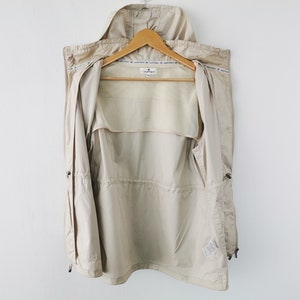 May include: A beige hooded jacket with a front zipper closure, featuring a drawstring waist, long sleeves, and a light-colored interior lining. The garment includes a small brand label at the neck and a repeating text pattern along the inner collar.