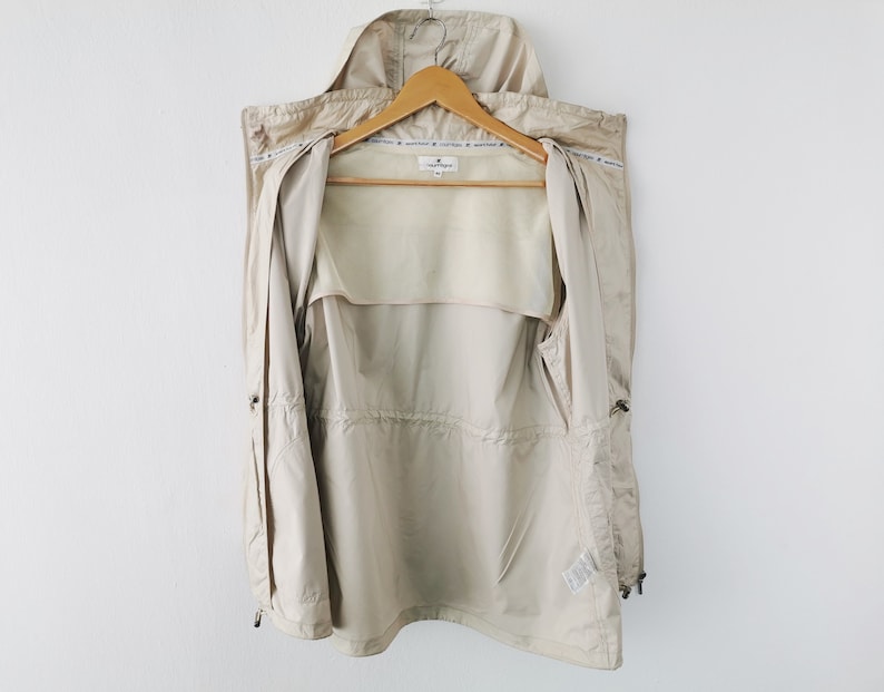 May include: A beige hooded jacket with a front zipper closure, featuring a drawstring waist, long sleeves, and a light-colored interior lining. The garment includes a small brand label at the neck and a repeating text pattern along the inner collar.